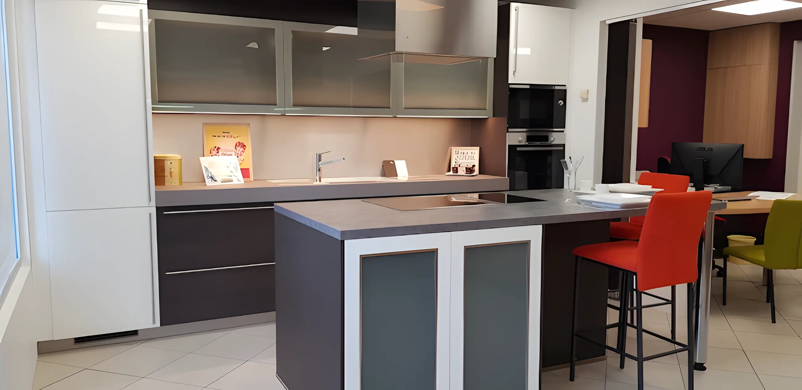 Cuisine exposée au showroom : îlot gris anthracite, meubles hauts vitrés et tabourets orange